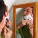 A man shaving with a shavette in a mirror.